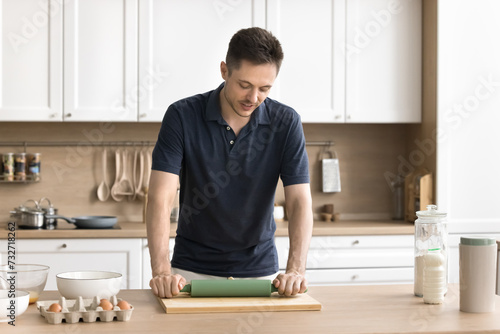 Handsome young man baking in home kitchen, rolling dough on wooden board, standing at table with fresh eggs, preparing pastry food, bakery dessert for holiday, festive dinner