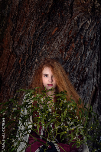 Portrait of a girl with red hair with a bouquet of mistletoe against the background of the textured bark of an old tree.