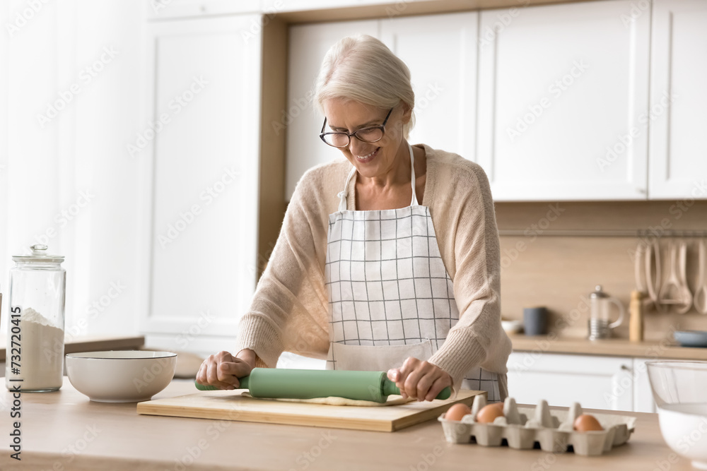 Positive mature baker woman wearing apron, rolling dough on home kitchen table with ingredients, preparing bakery meal for dinner, baking dessert, cake, pie, cookies, smiling Positive mature baker woman wearing apron, rolling dough on home kitchen table with ingredients, preparing bakery meal for dinner, baking dessert, cake, pie, cookies, smiling