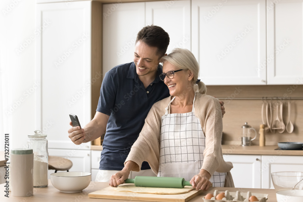 Happy young adult man and senior blonde mom baking in kitchen and talking on video call on smartphone, holding mobile phone, hugging, looking at gadget screen, smiling, laughing Happy young adult man and senior blonde mom baking in kitchen and talking on video call on smartphone, holding mobile phone, hugging, looking at gadget screen, smiling, laughing