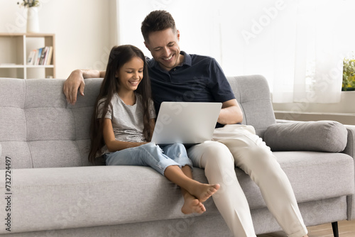 Cheerful young dad and pretty little schoolkid girl using Internet web service on laptop on home couch, watching funny movie, talking on video call, enjoying online communication, entertainment