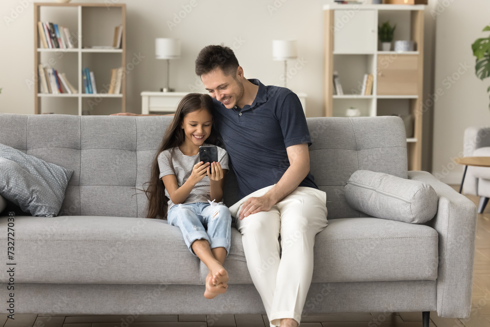 Happy positive father and little kid girl using smartphone for online communication, playing on digital device together, relaxing on comfortable home couch, enjoying leisure, Internet technology Happy positive father and little kid girl using smartphone for online communication, playing on digital device together, relaxing on comfortable home couch, enjoying leisure, Internet technology