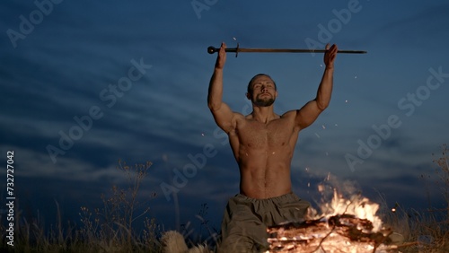 Swordsman heating the tip of his sword in a fire outdoors in open countryside at twilight before lifting the glowing weapon above his head and bringing it straight down in front of his bare chest. photo