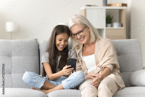 Cheerful granny and happy pre teen girl using funny application on mobile phone together, relaxing on home sofa with digital gadget. Kid showing grandma Internet service working on smartphone