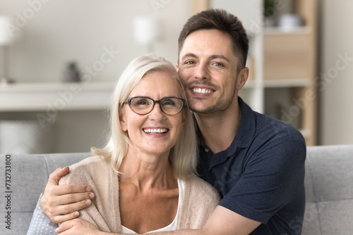 Cheerful caring adult son hugging happy blonde senior mother with love, support, looking at camera with perfect toothy smile, laughing. Elderly mother and grown male child headshot portrait photo