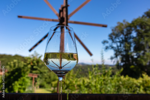 Wine tasting on vineyards and windmill klapotetz in Ehrenhausen an der Weinstrasse, Leibnitz, South Styria, Austria. Drinking glass of white wine on idyllic winery stretching over lush green hills photo