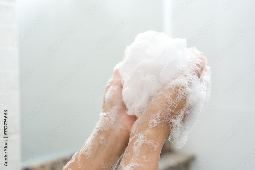 Fototapeta premium Hands woman millennial squeeze use foam taking shower. Female wash body with foam and stand under water shower. Lady washing hands with a sponge in the bathroom. Washing arms with a foamy sponge.