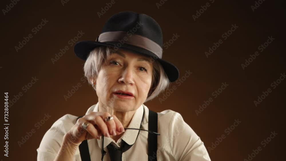 Beautiful woman over 60 wearing stylish glasses and smiling, dressed in retro attire with suspenders, necktie, and black hat, gracefully posing for the camera. Brown background. Camera 8K RAW. Beautiful woman over 60 wearing stylish glasses and smiling, dressed in retro attire with suspenders, necktie, and black hat, gracefully posing for the camera. Brown background. Camera 8K RAW.