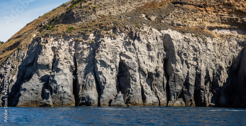 Weathered seaside rock face texture with parts of green and blue water. Aged volcanic stone wall surface background pattern with cracks and scratches. Banner. Ischia Island, Italy.