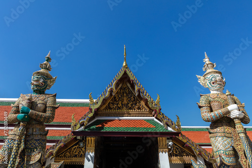 Colorful entrance to building in the Grand Palace, Bangkok, Thailand. Intricate gold decorations. Pair of Giant statues guarding the door. Blue sky behind. 
