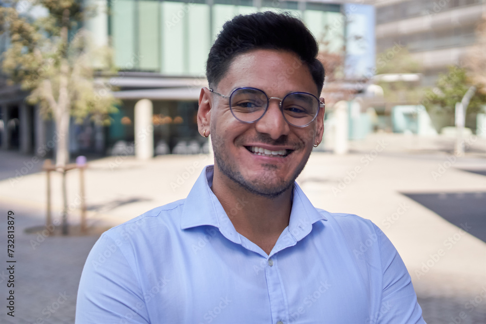 Obraz premium Portrait young adult Latin business man or commercial outside office center looking smiling at camera. Male freelance worker in shirt poses cheerful for job photo outdoors. Copy space staff people