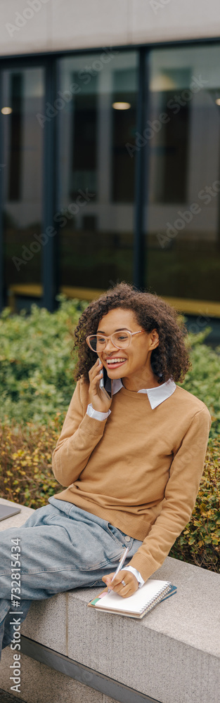 Smiling female freelancer talking phone and making notes while sitting on building background Smiling female freelancer talking phone and making notes while sitting on building background