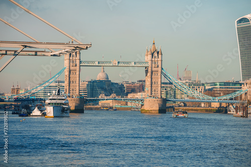 Sunny day at London's Tower Bridge with the River Thames in view, featuring boats and the distant dome of St Paul's Cathedral, highlighting the city's historic architecture. photo