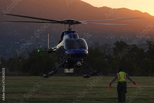 An aircraft marshaler guides an unmarked helicopter while landing in a field during the early morning, with mountains in the background.