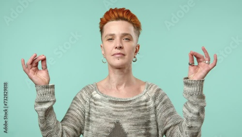 A composed lesbian woman with striking red hair and facial piercings lifts her hands in a meditative pose, seeking inner peace with her eyes closed against a soothing turquoise background. Camera RAW. photo
