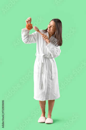 Young woman in bathrobe with loofah and massage brush on green background
