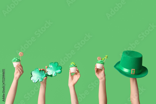 Female hands with tasty cupcakes, leprechaun hat and plastic eyeglasses for St. Patrick's Day on green background