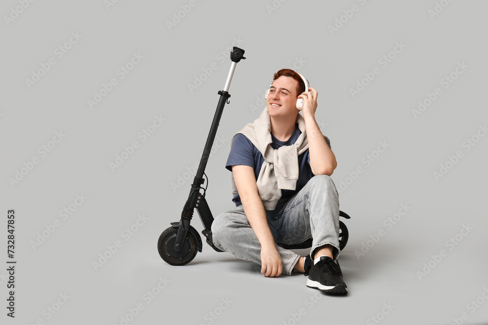 Young man in headphones with modern electric kick scooter sitting against grey background