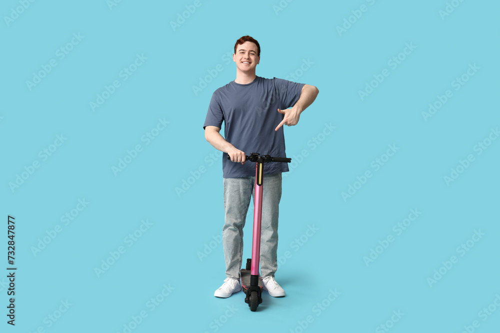 Young man pointing at modern electric kick scooter on blue background Young man pointing at modern electric kick scooter on blue background
