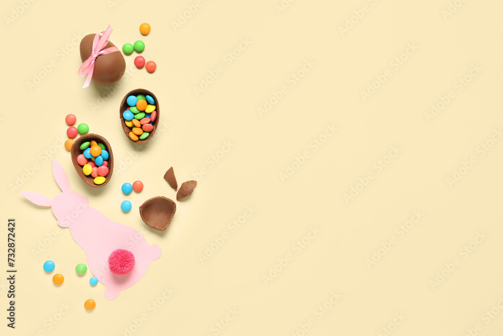 Chocolate Easter eggs with candies and paper bunny on yellow background