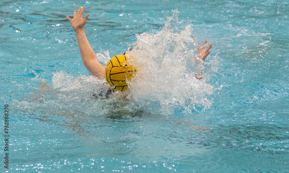 Two unrecognizable water polo players with obscured faces in pursuit of a yellow water polo ball while competing in a game in an indoor swimming pool