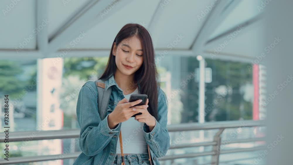 Young asian woman smiling, using mobile smart phone outdoor. Happy beautiful female wearing jeans jacket and holding smartphone at public city. Online technology and communication concept Young asian woman smiling, using mobile smart phone outdoor. Happy beautiful female wearing jeans jacket and holding smartphone at public city. Online technology and communication concept