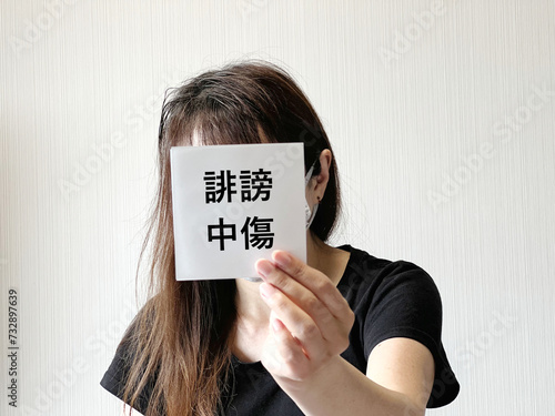 顔の前で誹謗中傷の文字入りの紙を片手で持つ女性