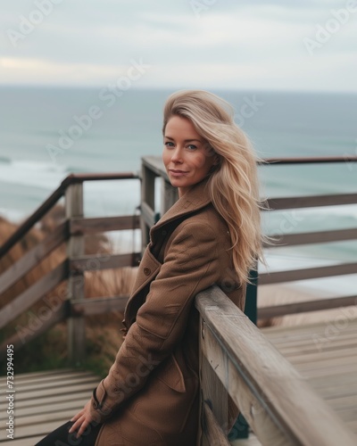 A poised Caucasian woman with flowing blonde hair, wearing a stylish brown coat, relaxes on a wooden seaside railing, embodying the casual grace of coastal autumn fashion © Oleg Kozlovskiy