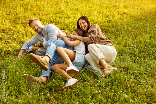 Happy young family of four lying on green grass and having fun in summer park. Smiling parents hugging and playing with kids boy and girl in nature enjoying time together. Family leisure concept.