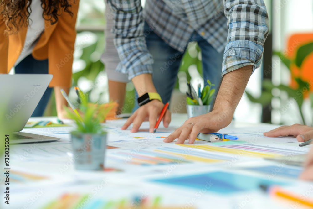 Fototapeta premium Close-up of a creative team's hands working together on colorful design plans spread across a table, with digital devices and stationery.