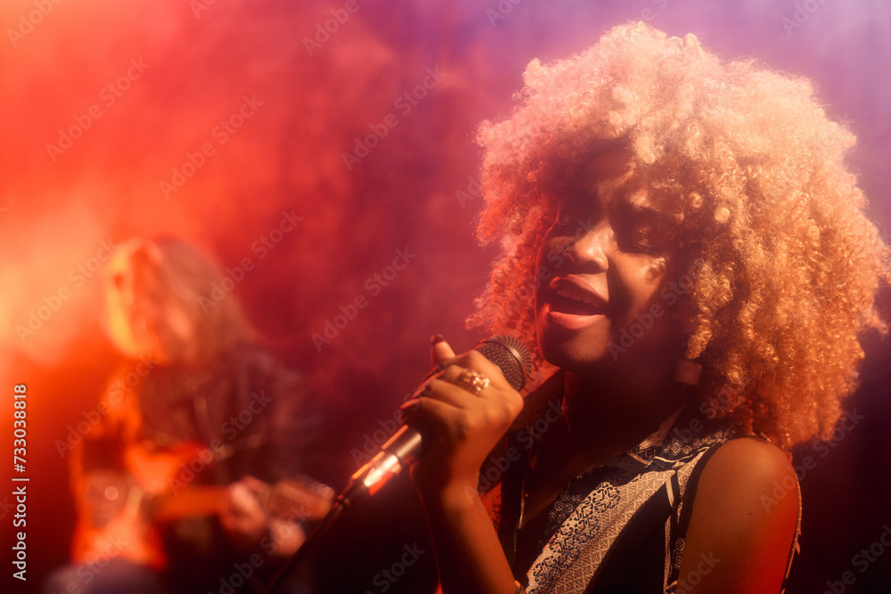 Naklejka premium Portrait of young Black woman singing passionately to microphone on stage in hazy neon lights copy space