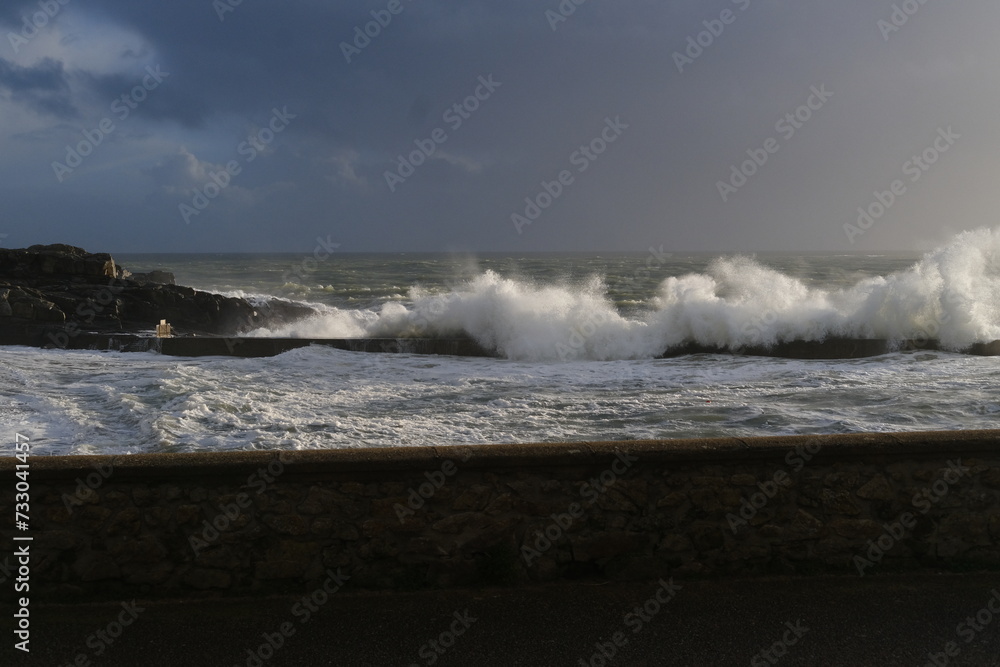 A huge wave is breaking on the jetty of a small French harbor on a stormy day. Batz-sur-Mer, France - February 10, 2024.