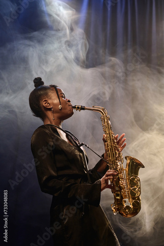 Vertical portrait of talented Black woman playing saxophone on stage during performance in jazz music club with smoke effect photo