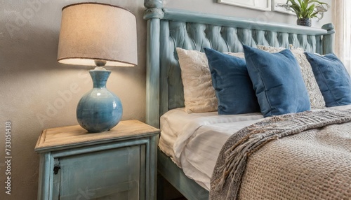 close up of blue ceramic lamp on nightstand near bed with beige fabric headboard and blue pillows and blanket french country provence interior design of modern bedroom