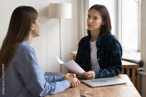 Serious young project manager woman in casual informal clothes talking to female business colleague at work table, using paper documents, holding resume, interviewing job candidate,