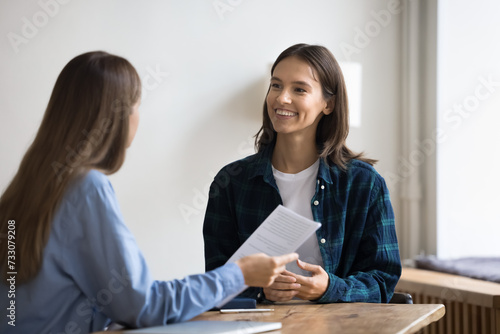 Happy young job candidate woman in informal clothes talking to employee on interview, talking to boss with toothy smile, meeting with colleague at work table, discussing paper resume photo