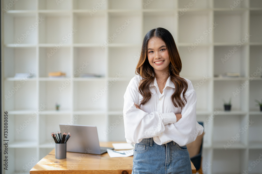 Smiling young woman standing confidently in a casual office environment, dressed in a white shirt and jeans.