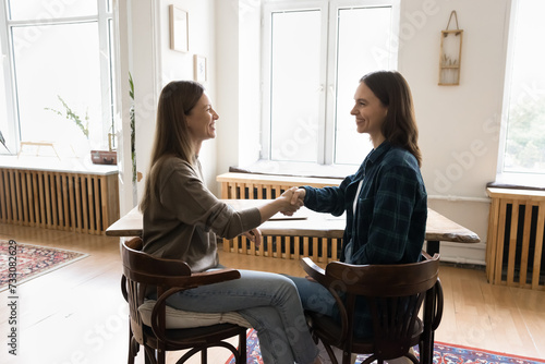 Two cheerful female entrepreneurs shaking hands at work table, meeting in office apartment for partnership, cooperation, teamwork, talking, smiling, laughing, ending negotiation in handshake