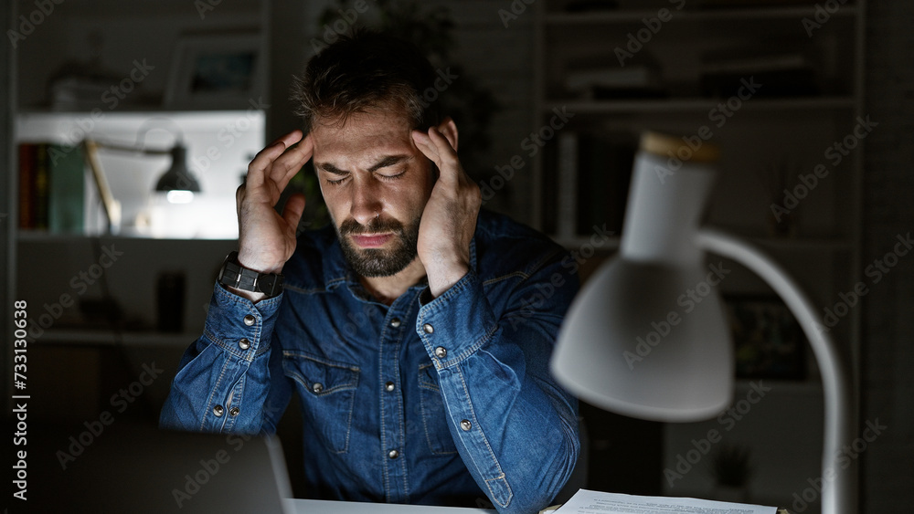 Overworked young hispanic man stressed from laptop work, grapples with office problem, dark night looms as pain from headache hurts, light from lamp reflected on his handsome beard Overworked young hispanic man stressed from laptop work, grapples with office problem, dark night looms as pain from headache hurts, light from lamp reflected on his handsome beard