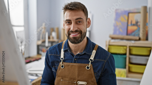 Portrait of a smiling young hispanic man, confidently sitting amid his art studio, brimming with paintings