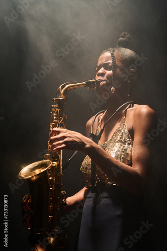 Side view portrait of young Black woman playing saxophone jazz music in spotlight