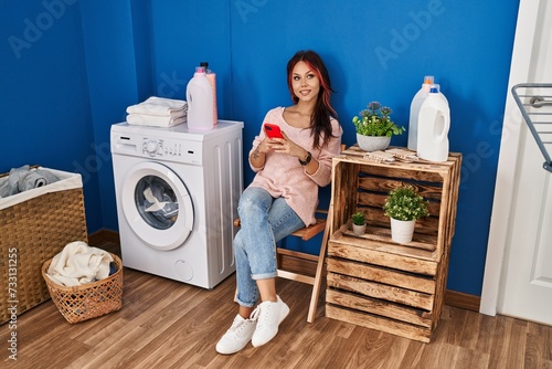 Young caucasian woman using smartphone waiting for washing machine at laundry room