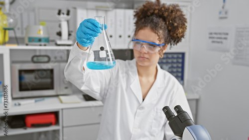 Obraz na plátně A young hispanic woman, with curly hair, examines a flask in a lab setting, illustrating science and healthcare
