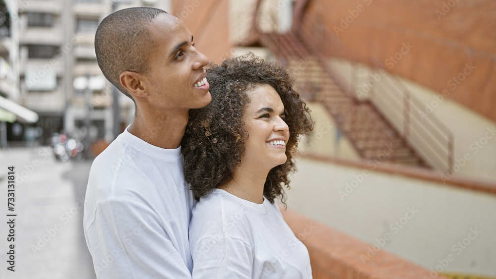 Beautiful couple, hugging and smiling, confidently look up at the sky on a buzzing city street. full of love and joy, they share this happy moment outdoors, basking in the sunlight. Beautiful couple, hugging and smiling, confidently look up at the sky on a buzzing city street. full of love and joy, they share this happy moment outdoors, basking in the sunlight.