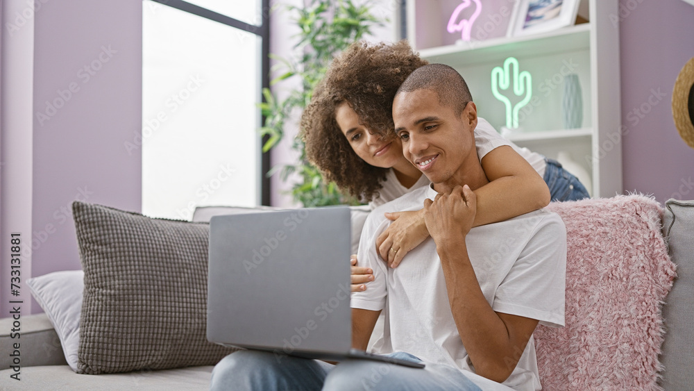 Naklejka premium Beautiful couple enjoying a fun, joyful time kissing and working on a laptop while sitting together on a casual sofa at home, their confident smiles reflecting love, positivity and a happy lifestyle.