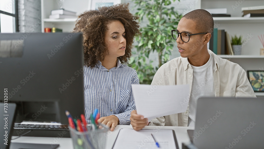Two office workers, man and woman, work together using computer and reading document at the office indoor Two office workers, man and woman, work together using computer and reading document at the office indoor