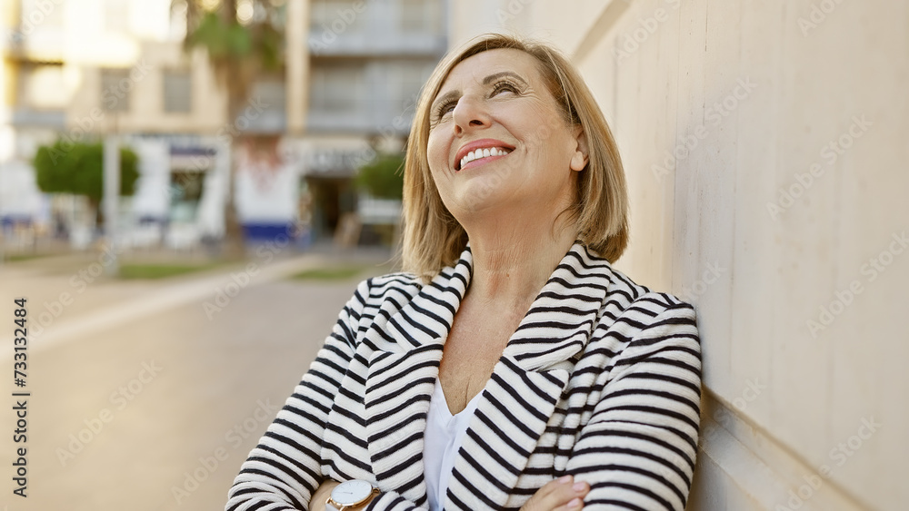 Mature caucasian woman standing outdoors in a city, smiling while leaning against a wall dressed in striped attire.