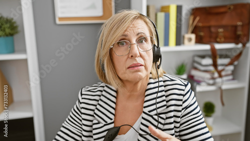A perplexed middle-aged woman with blonde hair wearing a headset in a modern office setting.