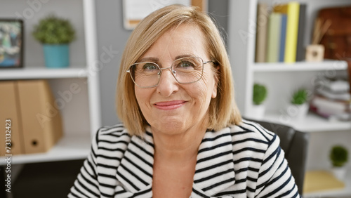 A smiling middle-aged woman with glasses in a striped blazer sits in a modern office