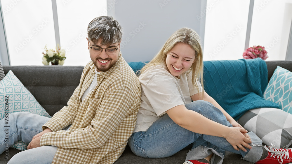 A happy man and woman sit back-to-back on a couch in a modern living room, sharing a moment of laughter together.